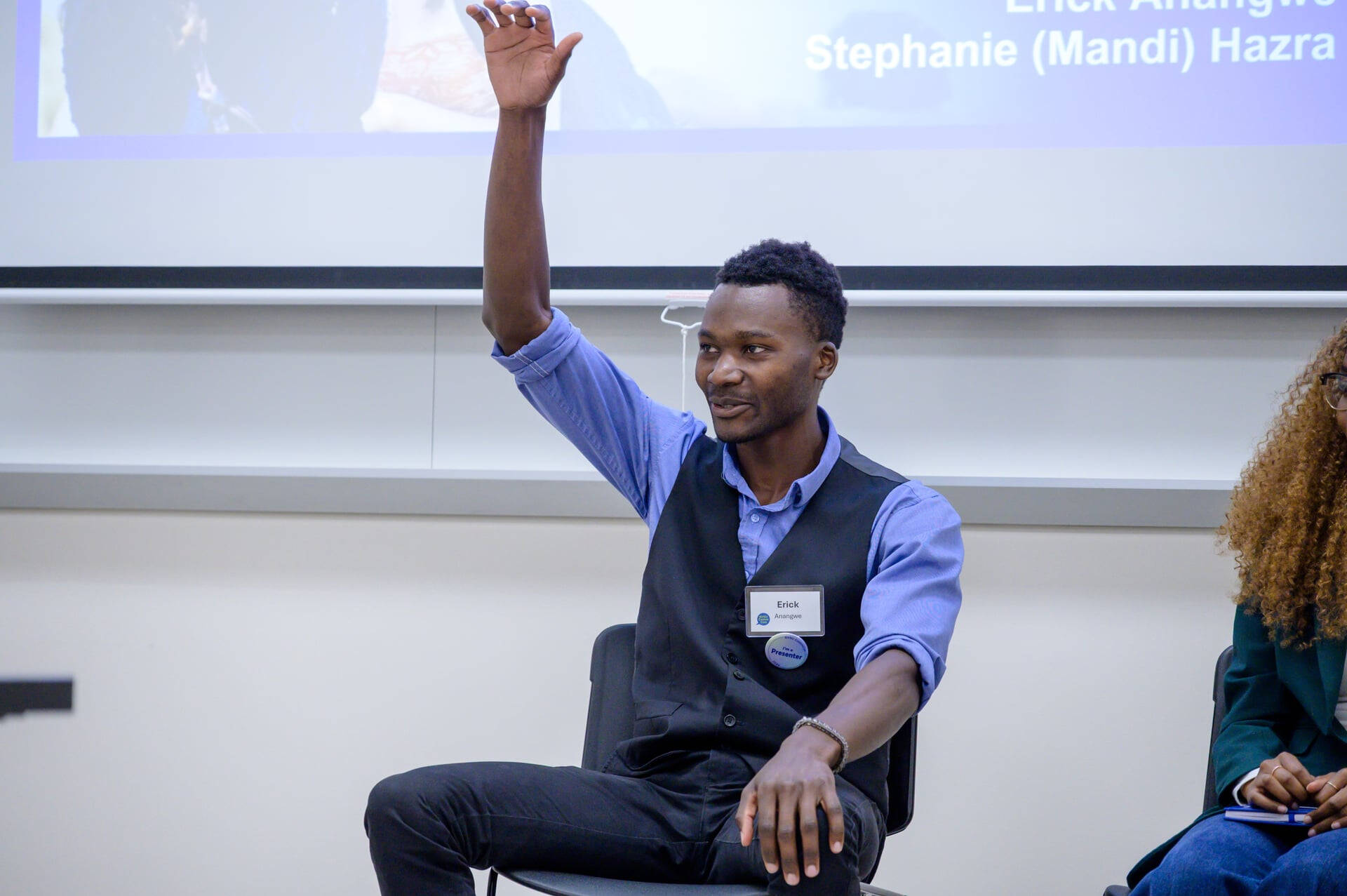 International student Erick Anangwe raises his hand during a panel discussion at GVSU CommCon. He is wearing a blue shirt and vest.